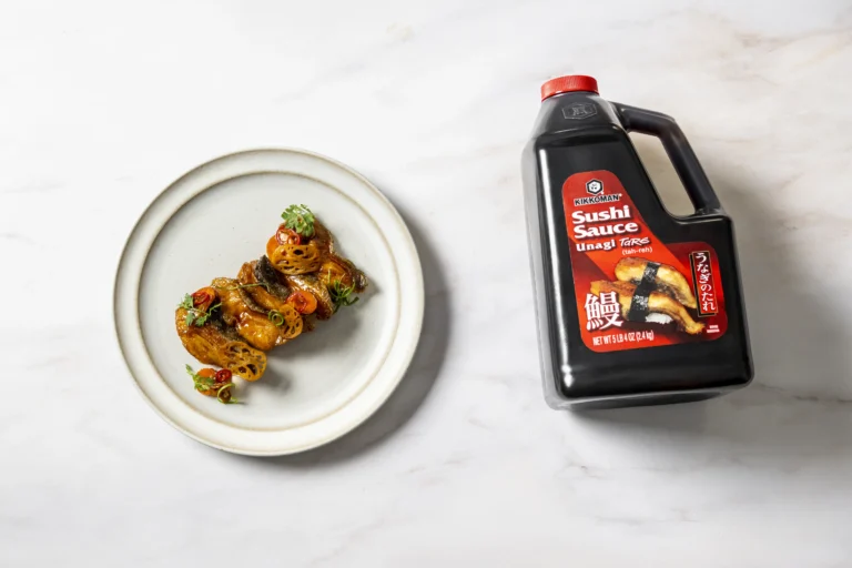 A plate of glazed eel garnished with herbs and cherry tomatoes is next to a large bottle of Kikkoman Sushi Sauce on a white surface.