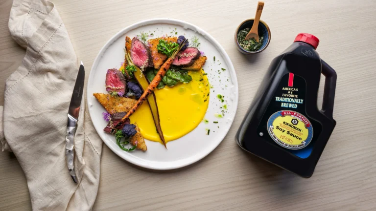 A plate of sliced steak with vegetables and yellow sauce sits next to a large bottle of traditionally brewed soy sauce on a light table.