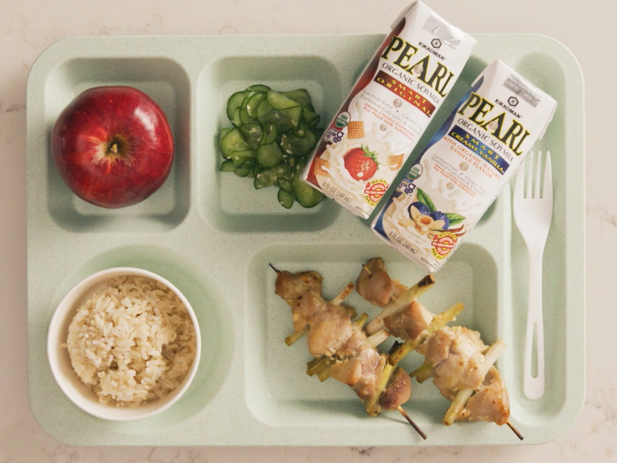 A cafeteria tray with an apple, sliced cucumbers, brown rice, grilled chicken skewers, a fork and spoon, and two cartons of PEARL organic soy milk.