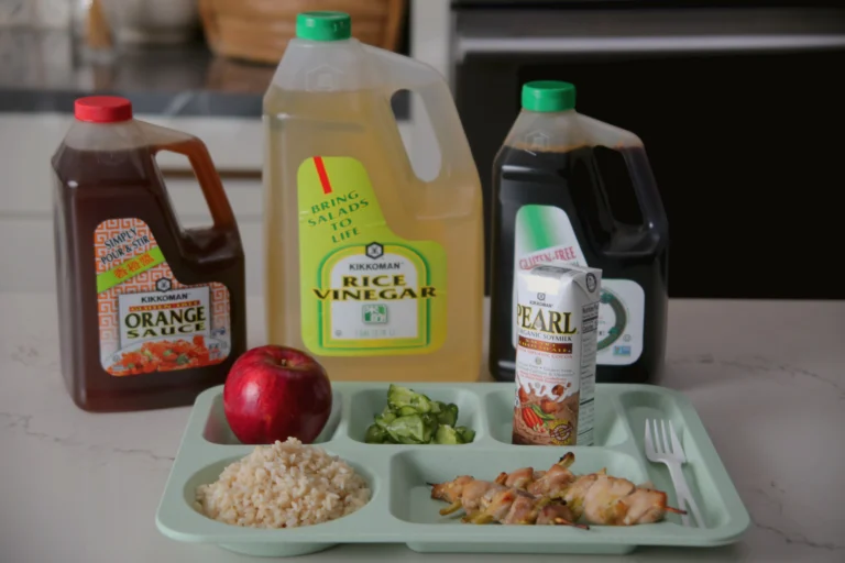 School lunch tray with brown rice, chicken skewers, cucumber salad, and an apple, in front of large bottles of orange sauce, rice vinegar, tamari, and a can of sparkling water.