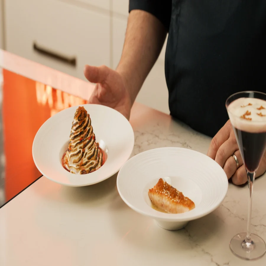 A person presents two plated dishes and a cocktail on a marble countertop; one dish features a baked Alaska dessert, the other a small piece of cooked fish.