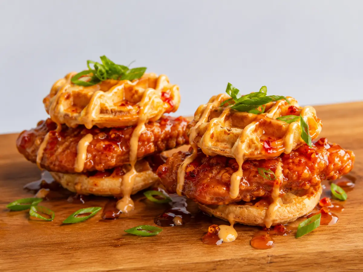 Two stacks of fried chicken and waffles topped with creamy sauce and sliced green onions on a wooden board.