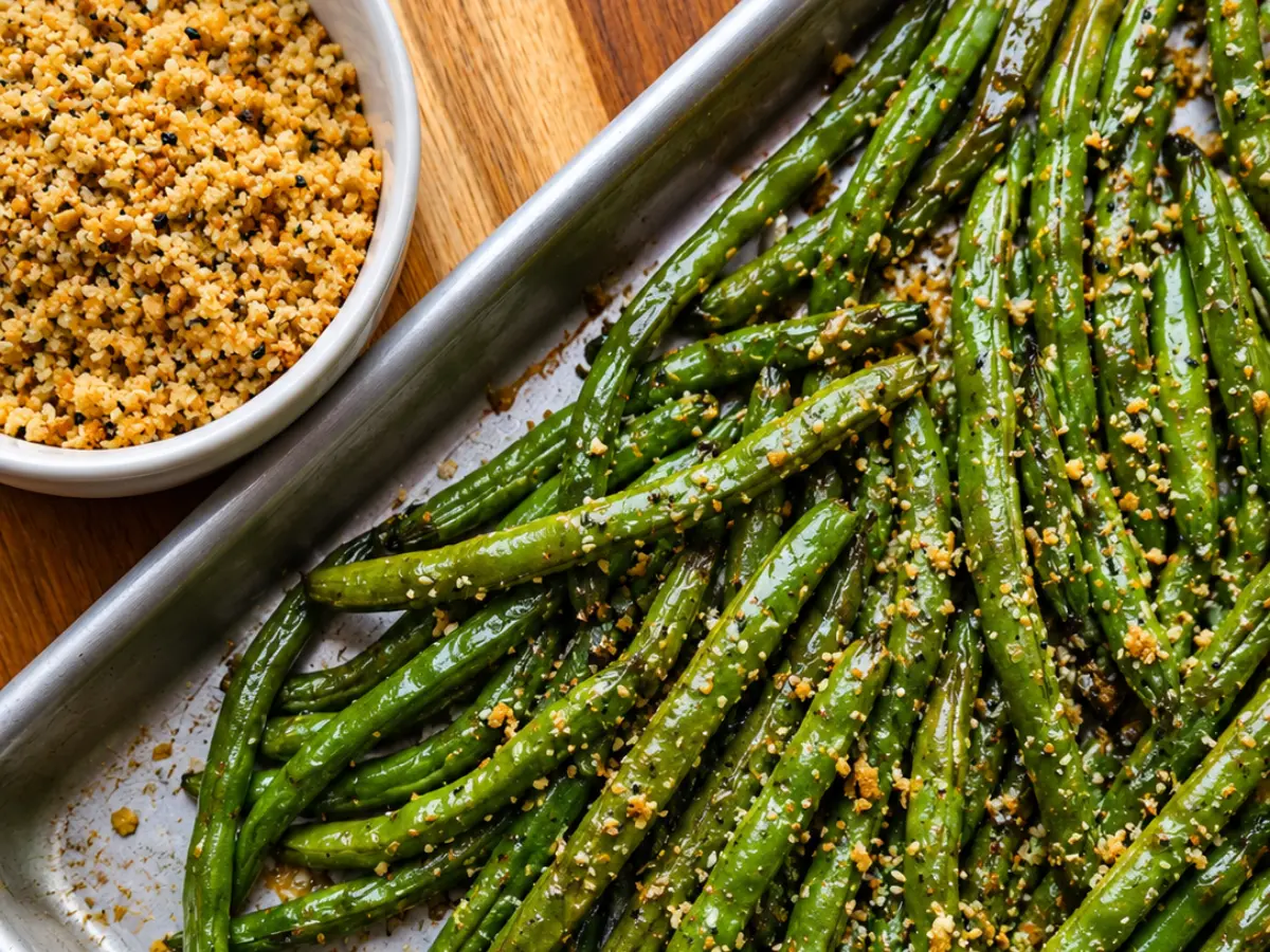 Roasted green beans topped with seasoned breadcrumbs on a baking sheet, next to a bowl of extra breadcrumbs on a wooden surface.