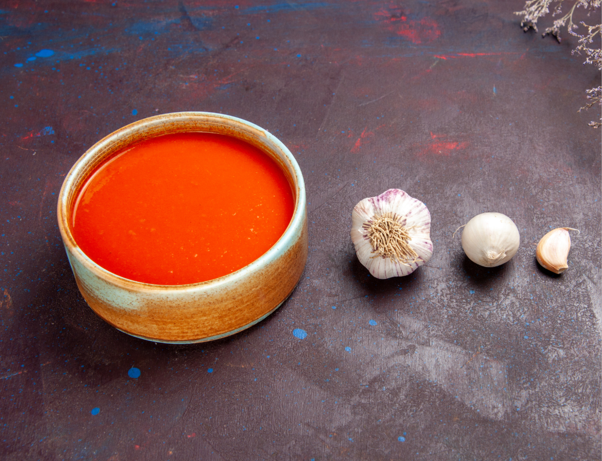 A bowl of red tomato sauce sits next to a garlic bulb, a white onion, and a garlic clove on a dark surface.