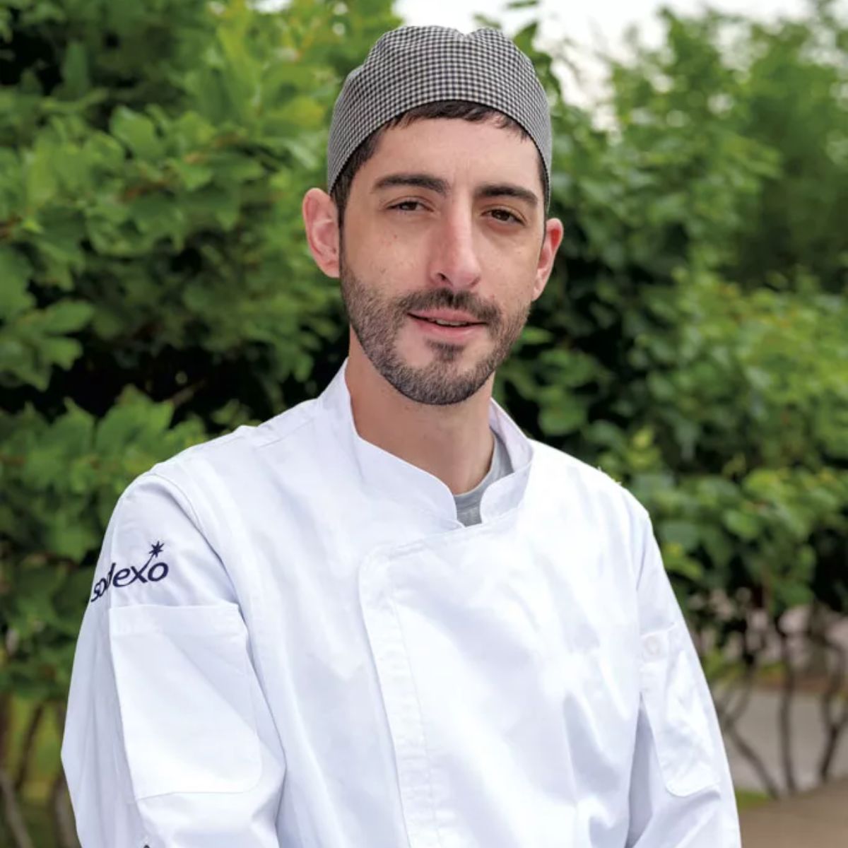 A man wearing a white chef's coat and a checkered hat stands outdoors in front of green foliage, looking at the camera.