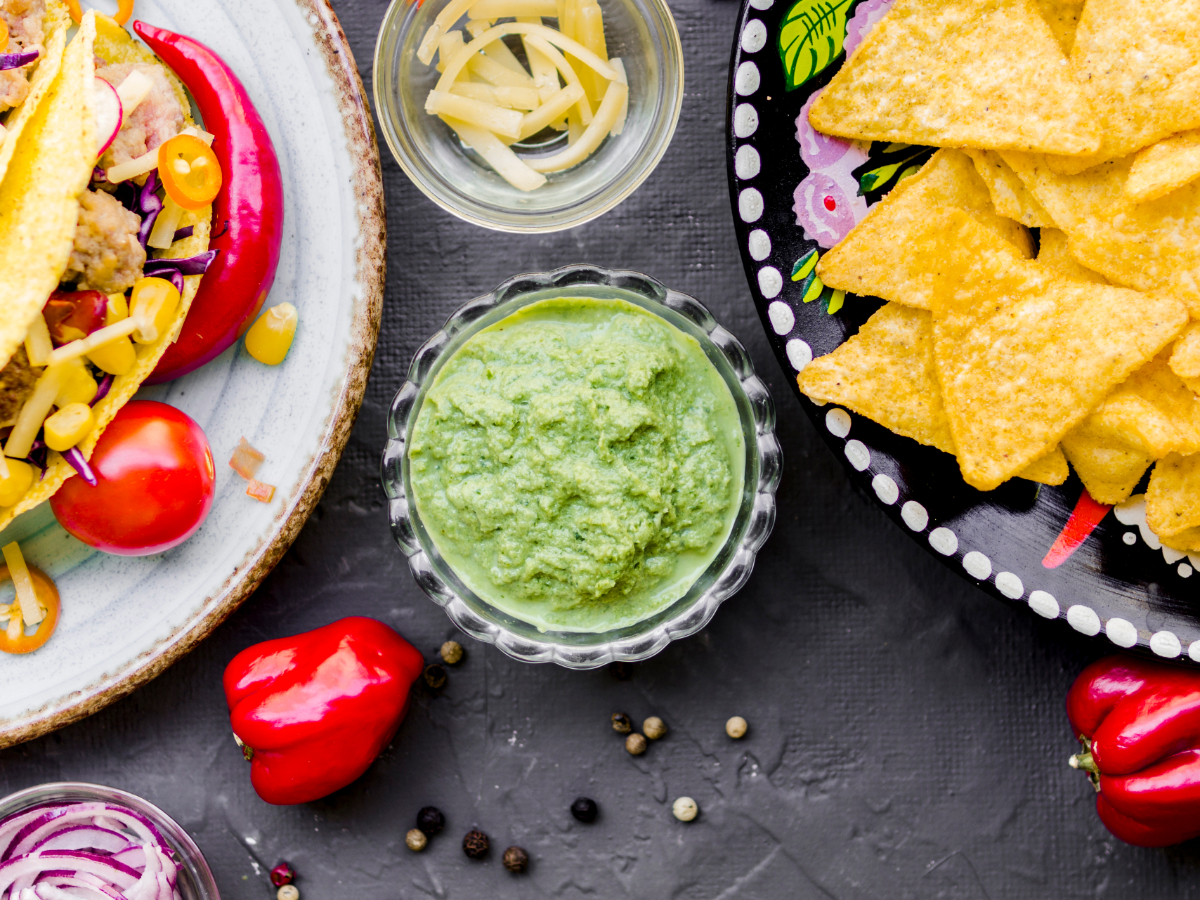 A bowl of green dip, a plate of yellow tortilla chips, sliced cheese, and a plate of topped tacos with fresh peppers on a dark surface.