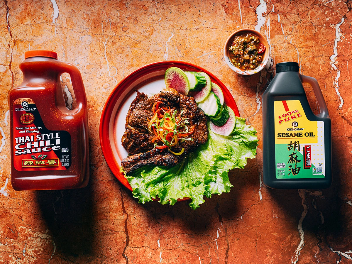 A plate with grilled meat and vegetables is flanked by a jug of Thai style chili sauce and a bottle of sesame oil; a small bowl of sauce sits above the plate on a reddish marble surface.