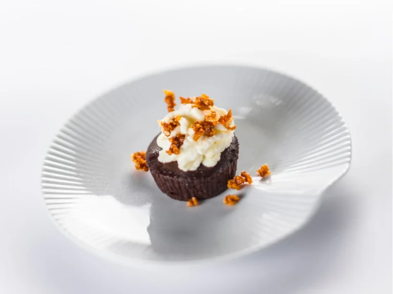 A chocolate dessert topped with whipped cream and caramelized sugar pieces sits on a white, shell-shaped plate against a white background.