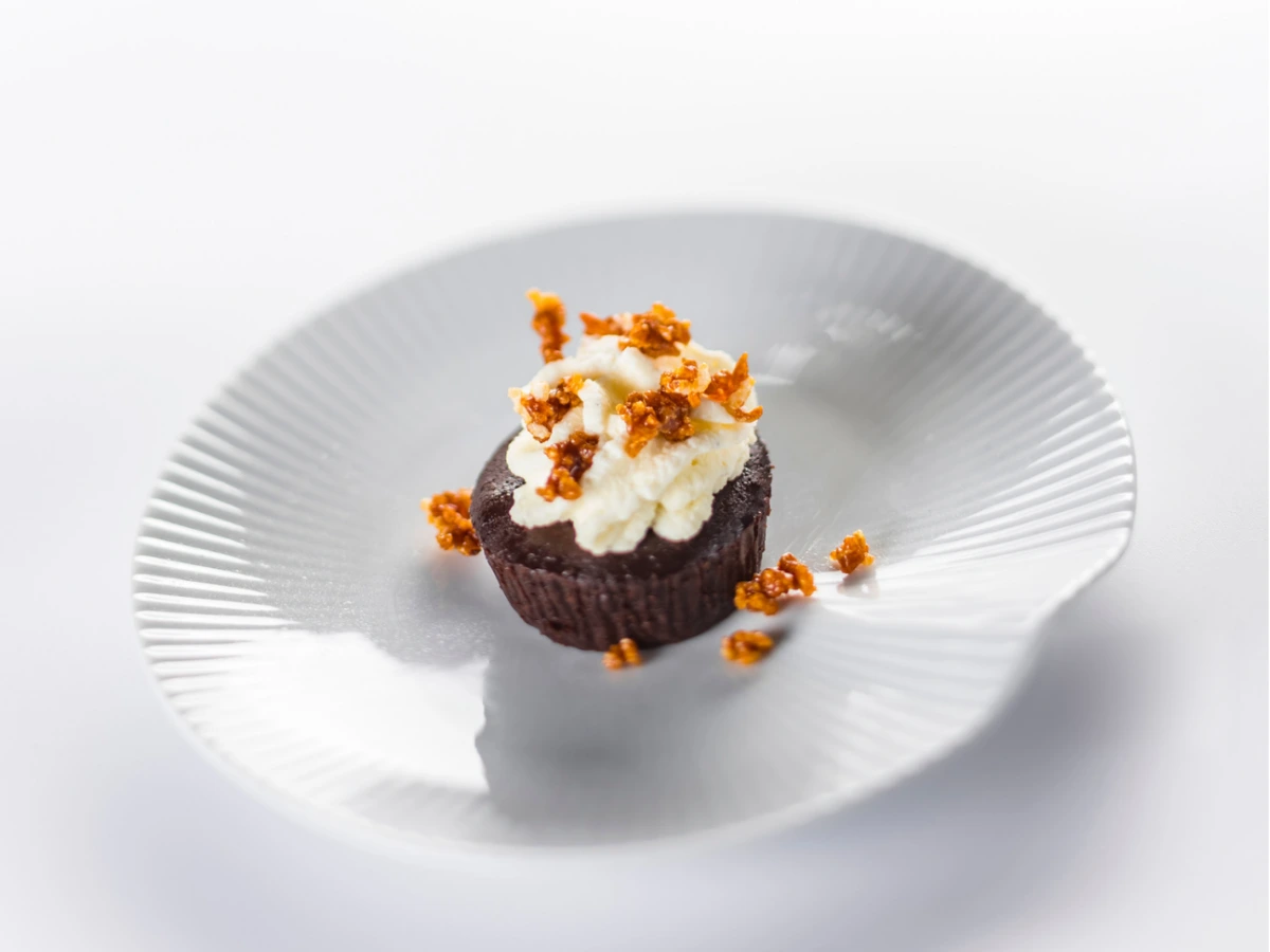 A chocolate dessert topped with whipped cream and caramelized sugar pieces sits on a white, shell-shaped plate against a white background.