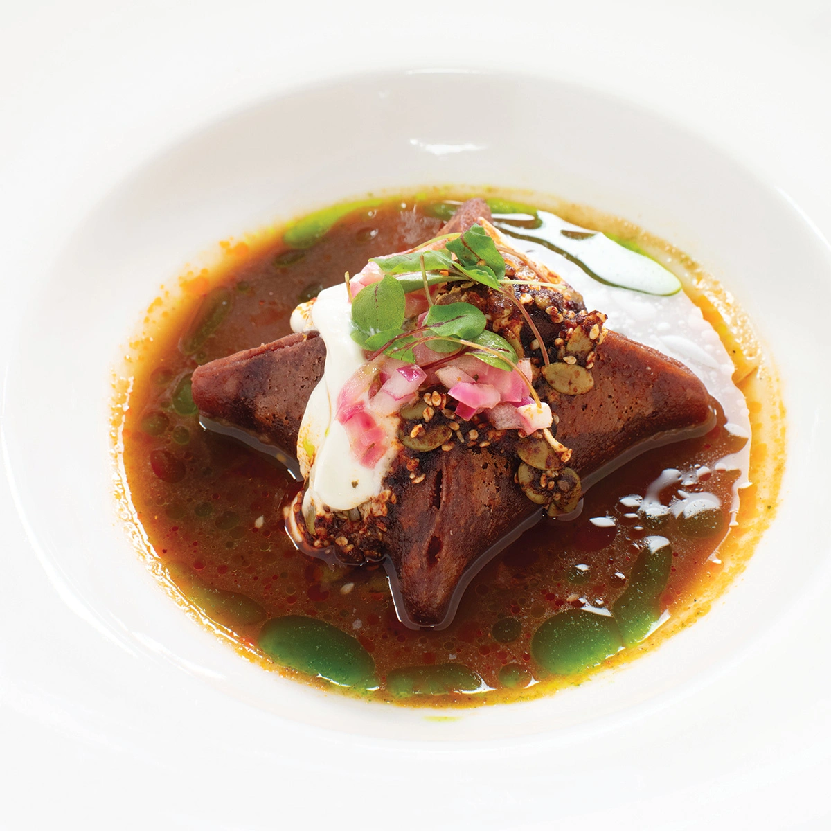 A star-shaped food item in broth, topped with cream, mustard seeds, chopped onions, and microgreens, served on a white plate.