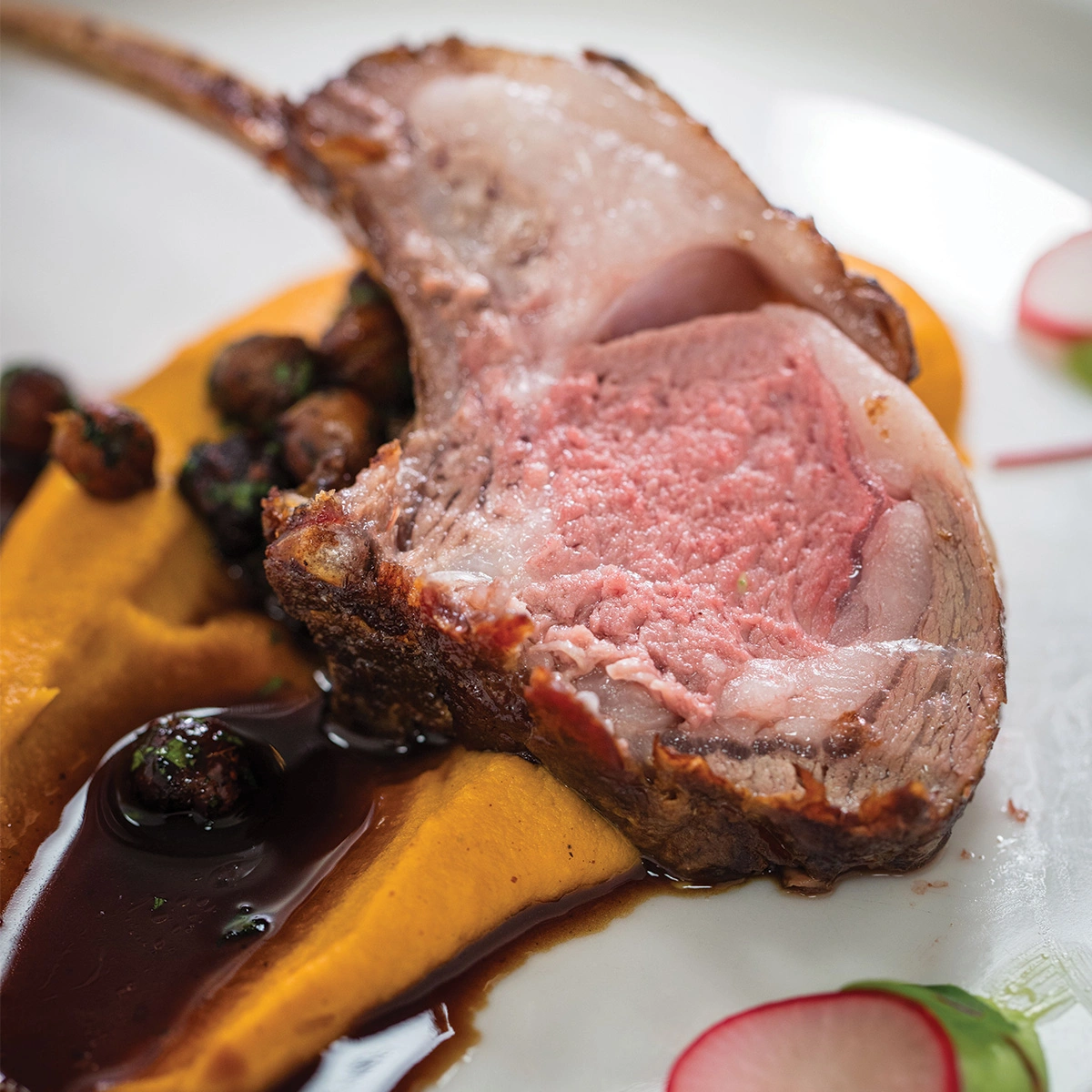 A cooked lamb chop rests on a swirl of orange puree with dark sauce and small garnishes nearby on a white plate.