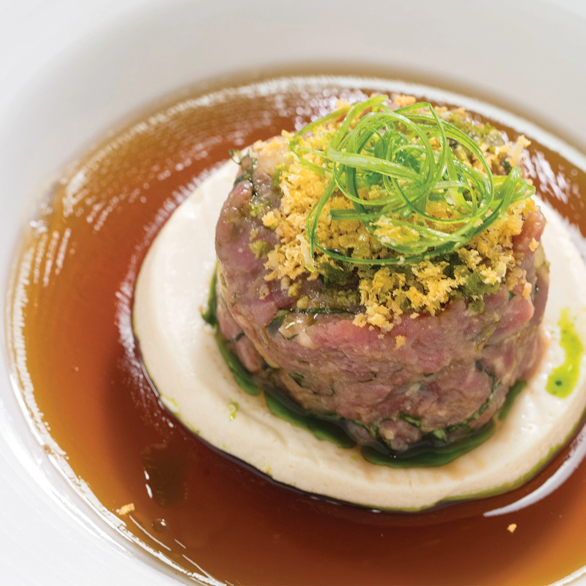 A round portion of beef tartare sits on a white sauce, topped with green onions and crispy crumbs, surrounded by a brown broth in a white bowl.