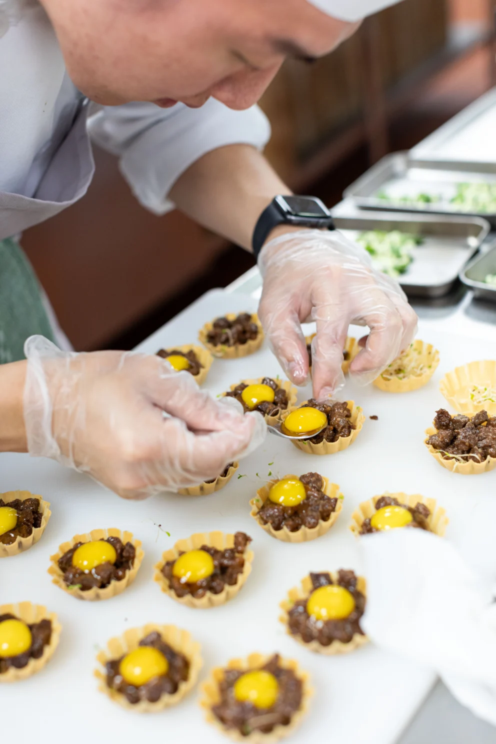 A chef wearing gloves arranges mini tartlets with minced meat and raw egg yolks on a white countertop.