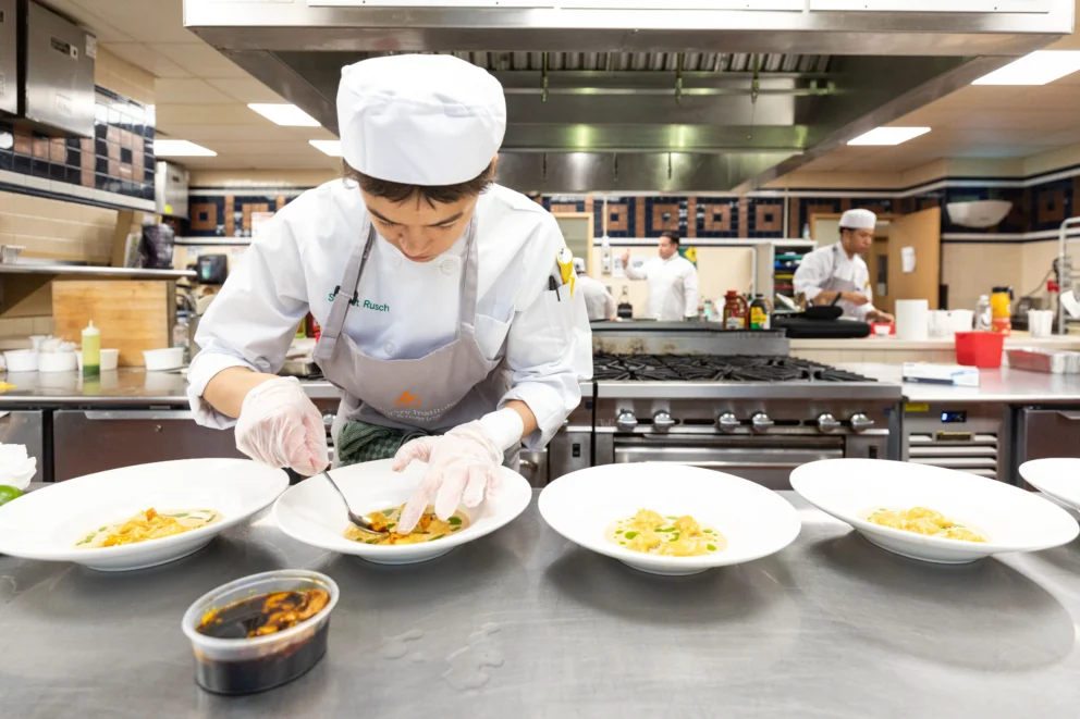A chef in uniform prepares pasta dishes in a commercial kitchen, carefully arranging food on plates lined up on a stainless steel counter.