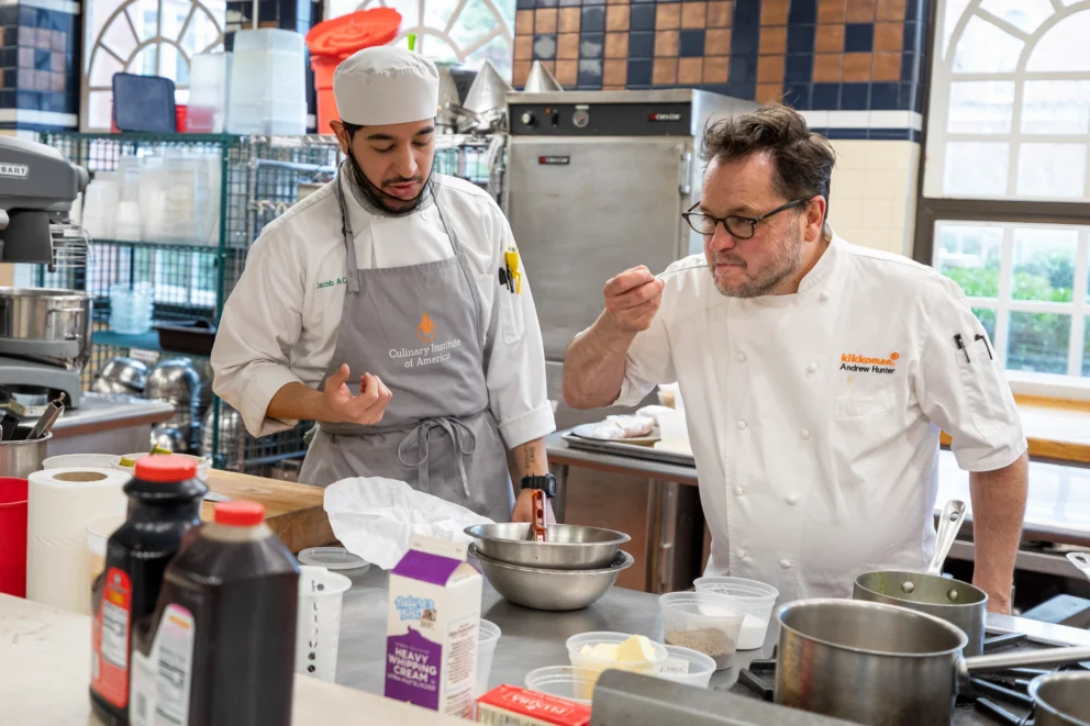 Two chefs in a commercial kitchen taste food from a metal bowl at a prep table surrounded by cooking ingredients and utensils.