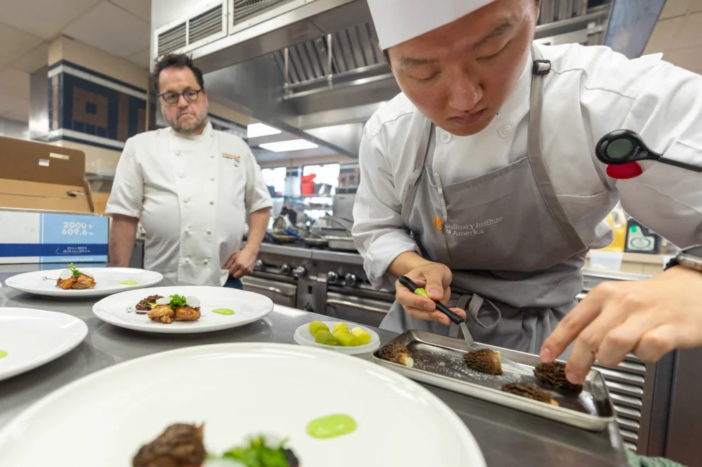 A chef in uniform plates food with tweezers while another chef watches nearby in a professional kitchen setting.