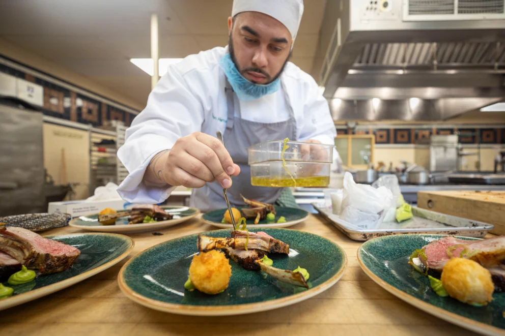 A chef in a white uniform and hat carefully plates food in a professional kitchen, adding sauce to dishes arranged on green plates.