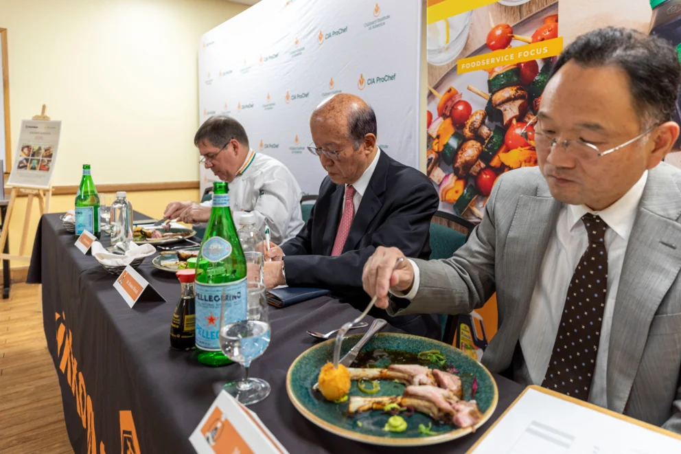 Three men in suits sit at a table, tasting and judging plated food dishes at a culinary competition event.