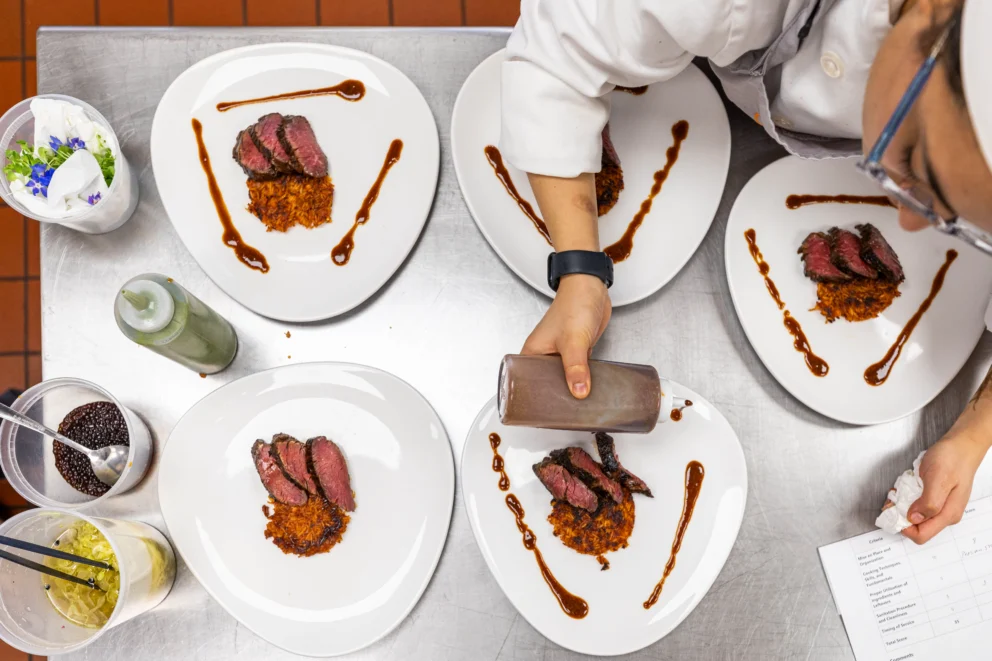 Chef plating five dishes with sliced steak on a bed of shredded vegetables, adding sauce in decorative lines on each plate, in a professional kitchen setting.