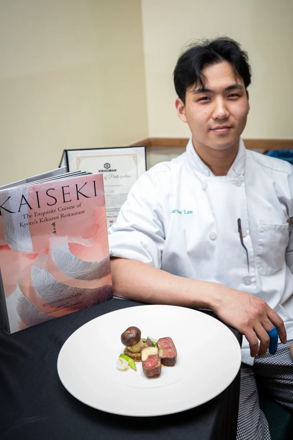 A chef in a white uniform sits at a table with a Kaiseki cookbook and a white plate featuring a small, artfully arranged dish of meat, mushroom, and vegetables.