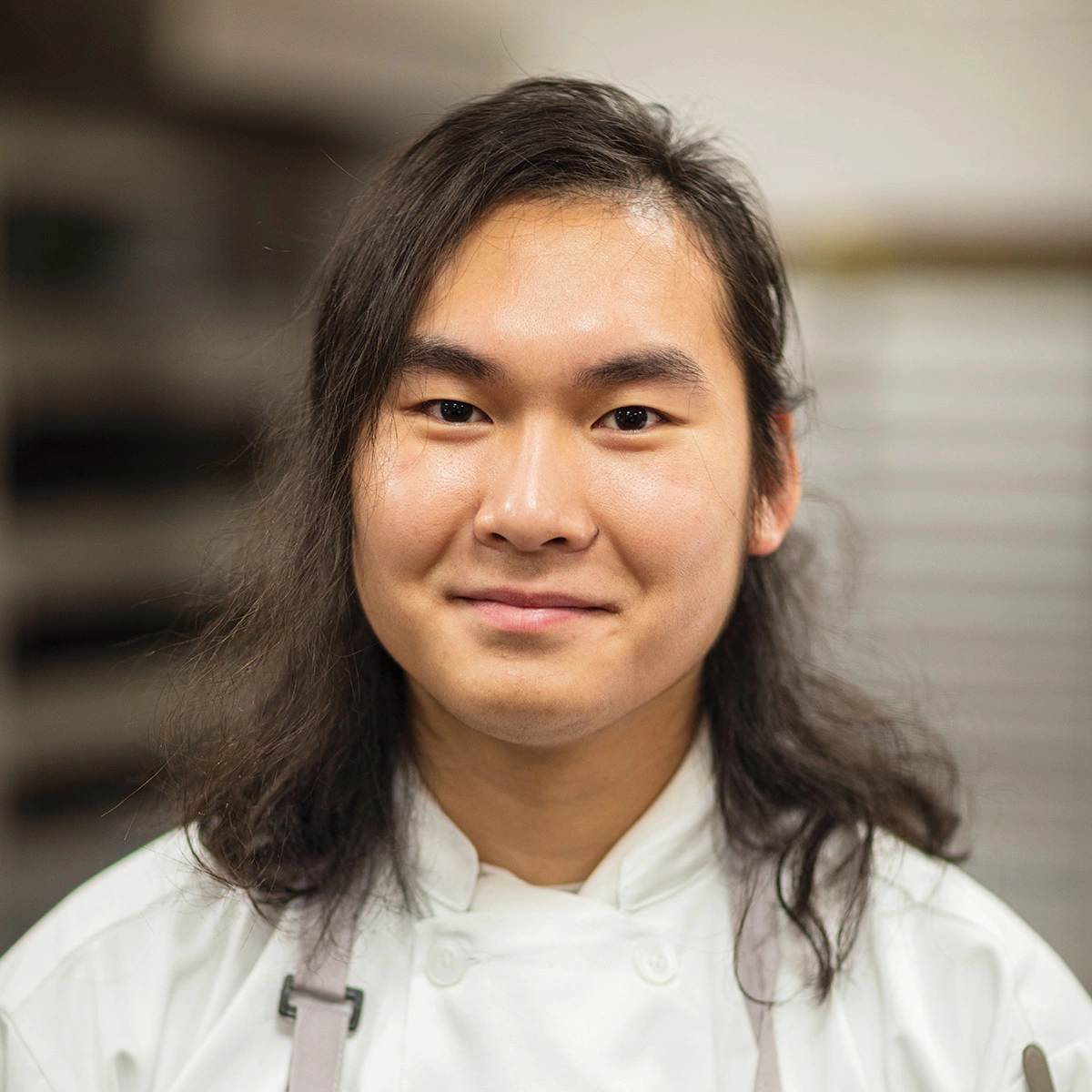 A person with long dark hair wearing a white chef’s coat and apron stands indoors, looking at the camera with a slight smile.