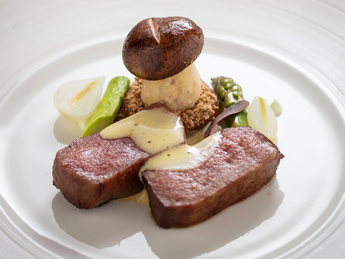 Two slices of cooked meat with creamy sauce, asparagus, mushroom, onion, and a crumb mixture arranged on a white plate.