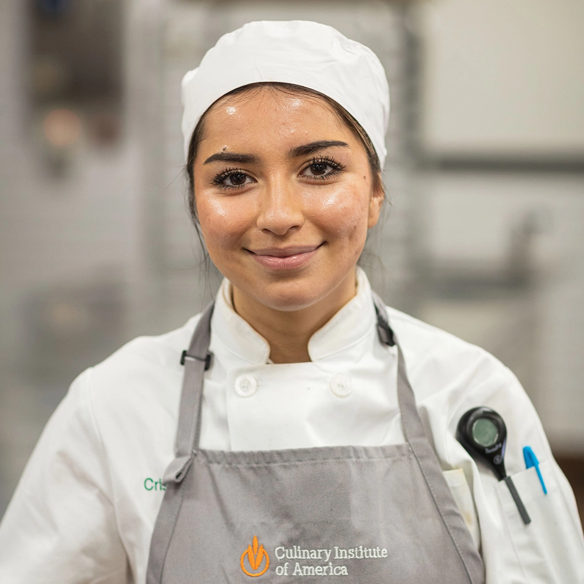 Young chef wearing a white hat, white chef’s coat, and gray apron stands in a kitchen. The apron reads “Culinary Institute of America.” She is smiling and looking at the camera.