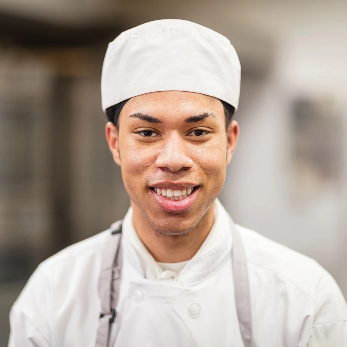 A person wearing a white chef’s uniform and hat is smiling while standing indoors in a kitchen setting.