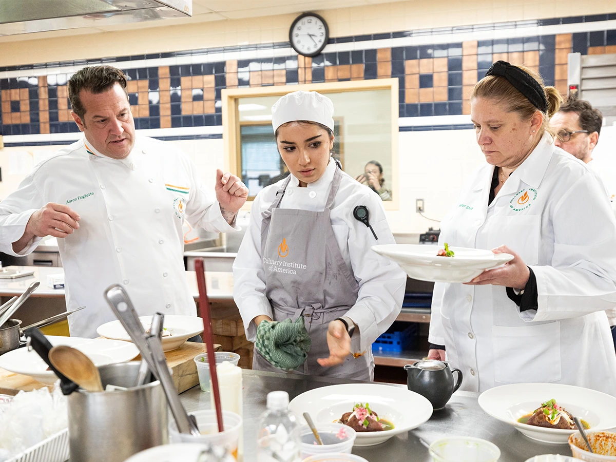 Three chefs in a commercial kitchen examine plated dishes, with one chef in the center holding a towel and listening attentively to the others.