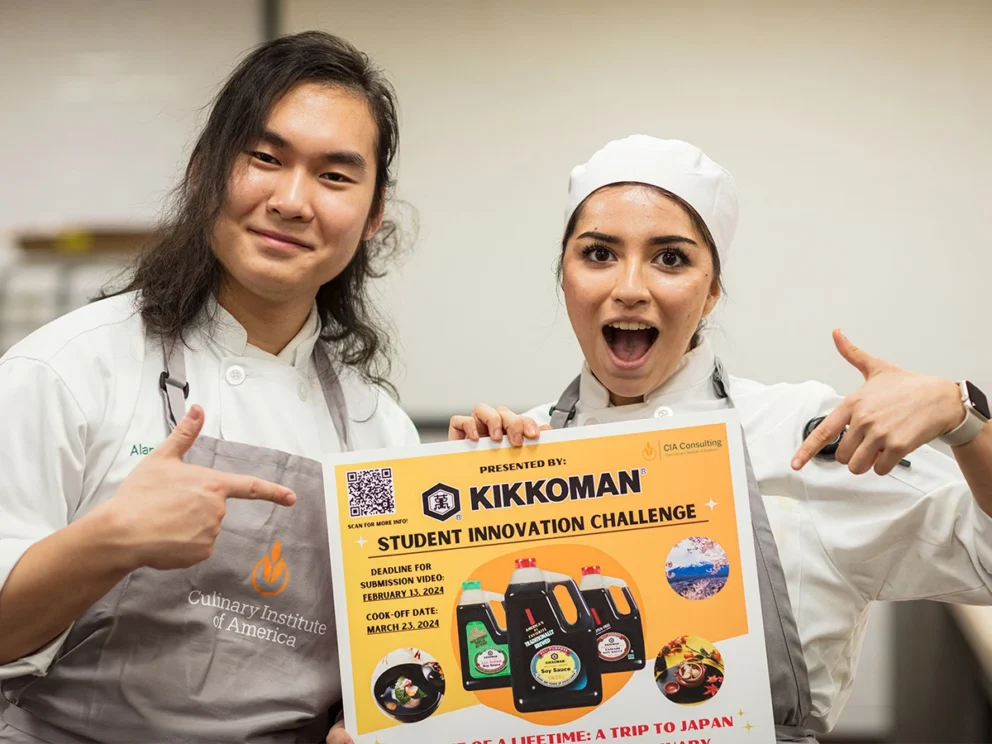 Two culinary students hold and point to a Kikkoman Student Innovation Challenge poster, promoting a contest with a chance to win a trip to Japan.