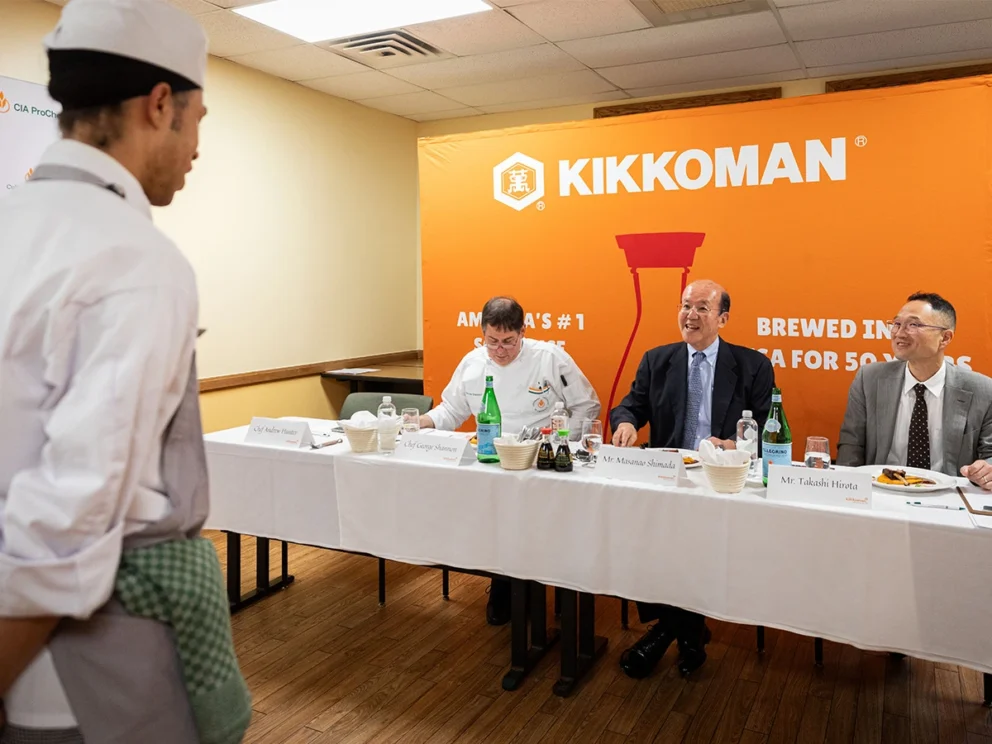 A chef stands before three seated judges at a table in front of a Kikkoman banner in a well-lit room with wooden floors.