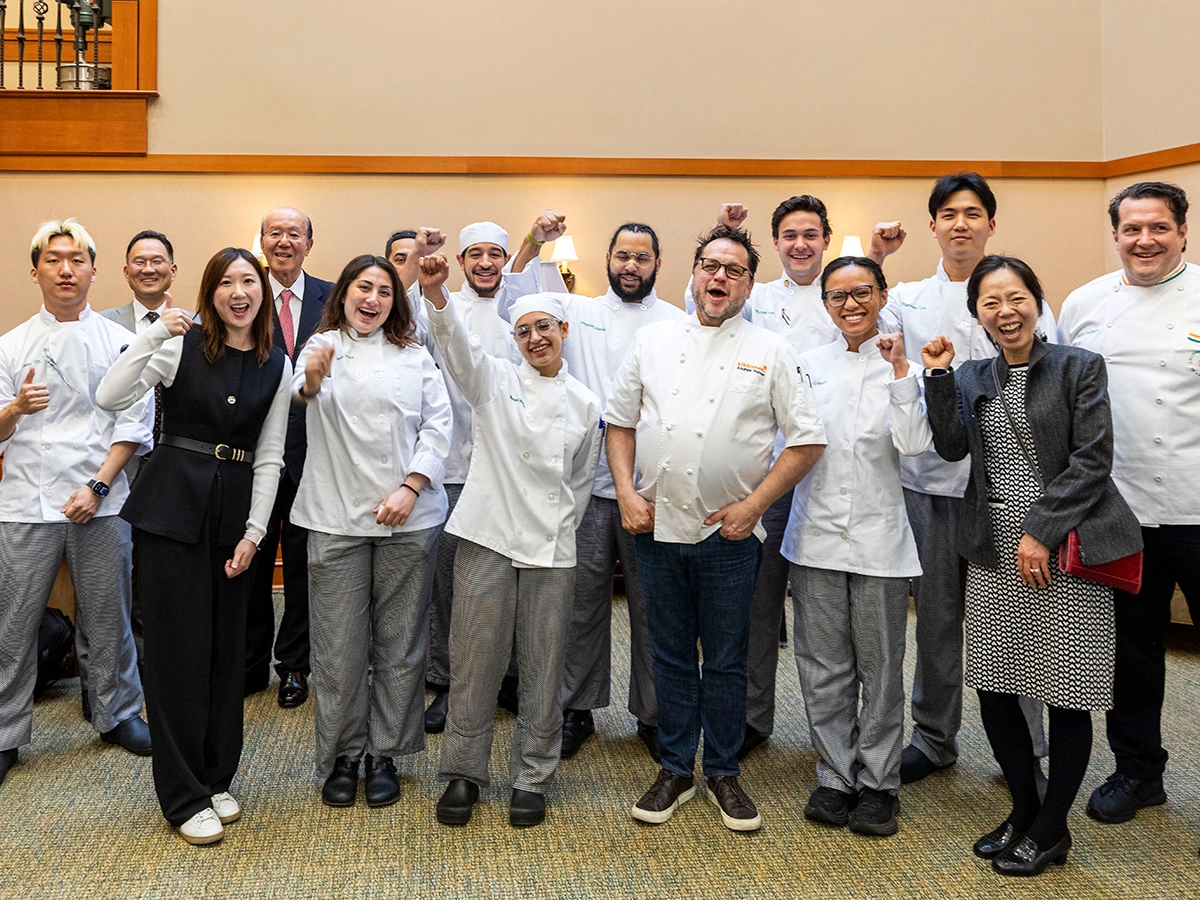 A group of chefs and individuals pose indoors, smiling and raising their fists, dressed in chef uniforms and business attire.