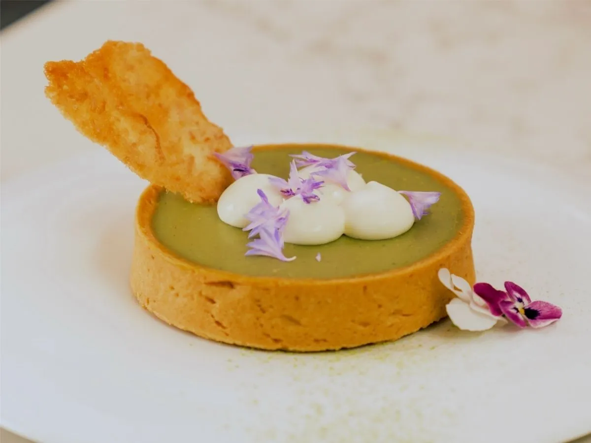 A matcha tart with whipped cream, edible flowers, and a cookie garnish is served on a white plate.