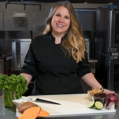 A woman in a black chef coat stands in a commercial kitchen behind a counter with cilantro, sweet potatoes, lime, onion, nuts, and a knife.