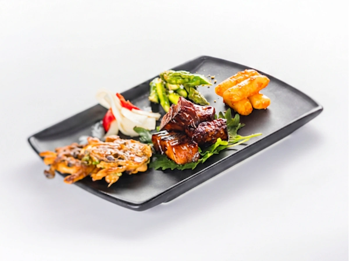 Rectangular black plate with assorted foods, including grilled meat, fried vegetables, gnocchi, asparagus, and pickled onions, arranged neatly on a white background.