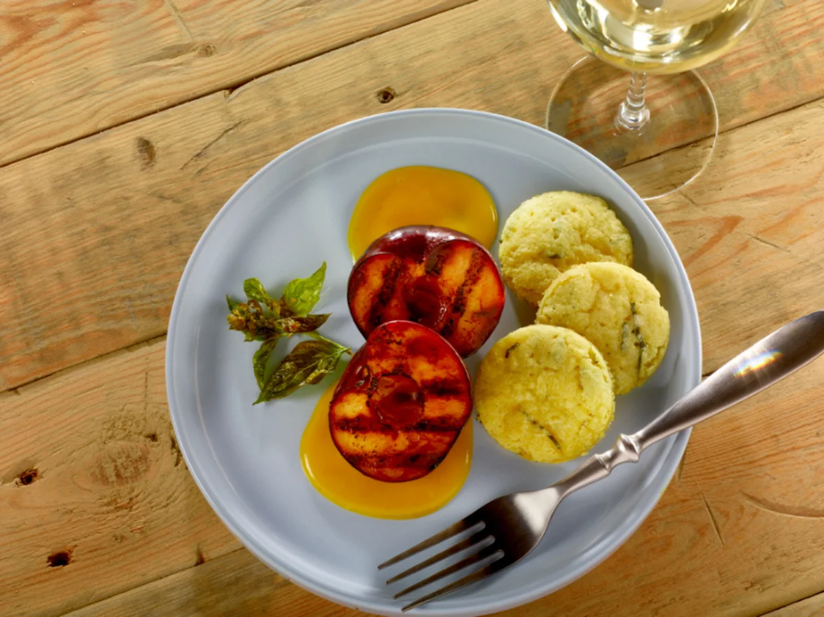 A plate with two grilled peach halves, yellow sauce, two biscuit rounds, herbs, and a fork, on a wooden table next to a glass of white wine.