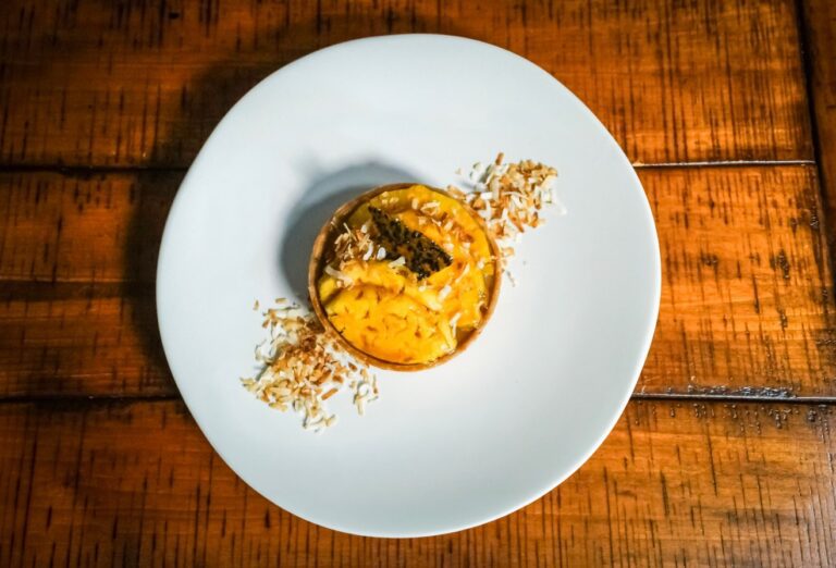 A small tart topped with shredded coconut and dark garnish is served on a white plate with toasted coconut flakes, placed on a wooden table.