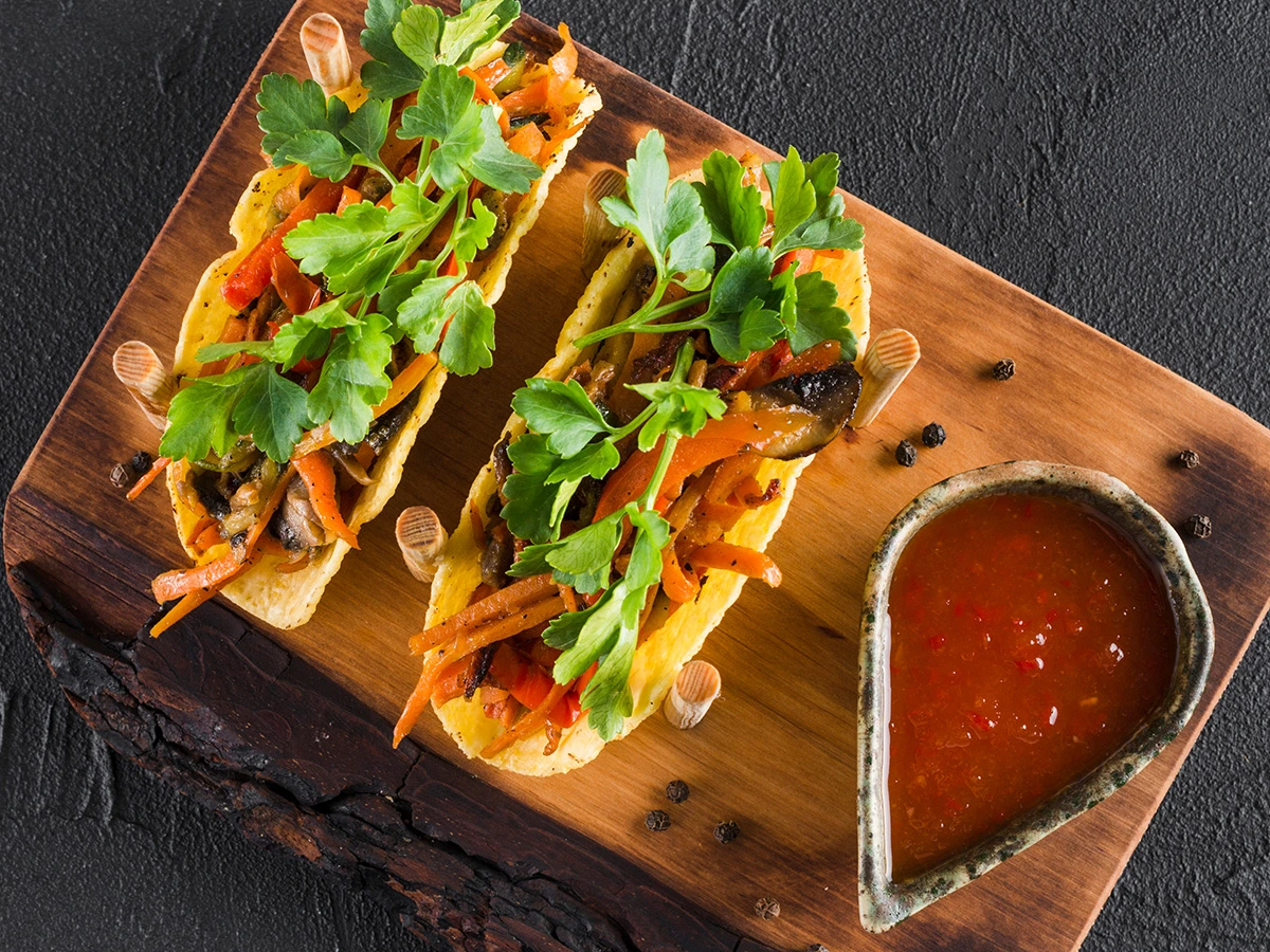 Two tacos filled with vegetables and fresh herbs on a wooden board, served with a small dish of red sauce on the side.