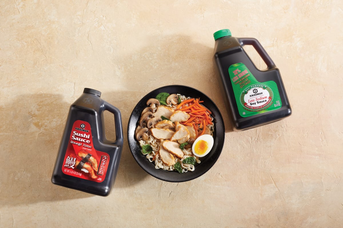 A bowl of ramen with chicken, vegetables, and egg is placed between large containers of sushi sauce and low sodium soy sauce on a beige surface.