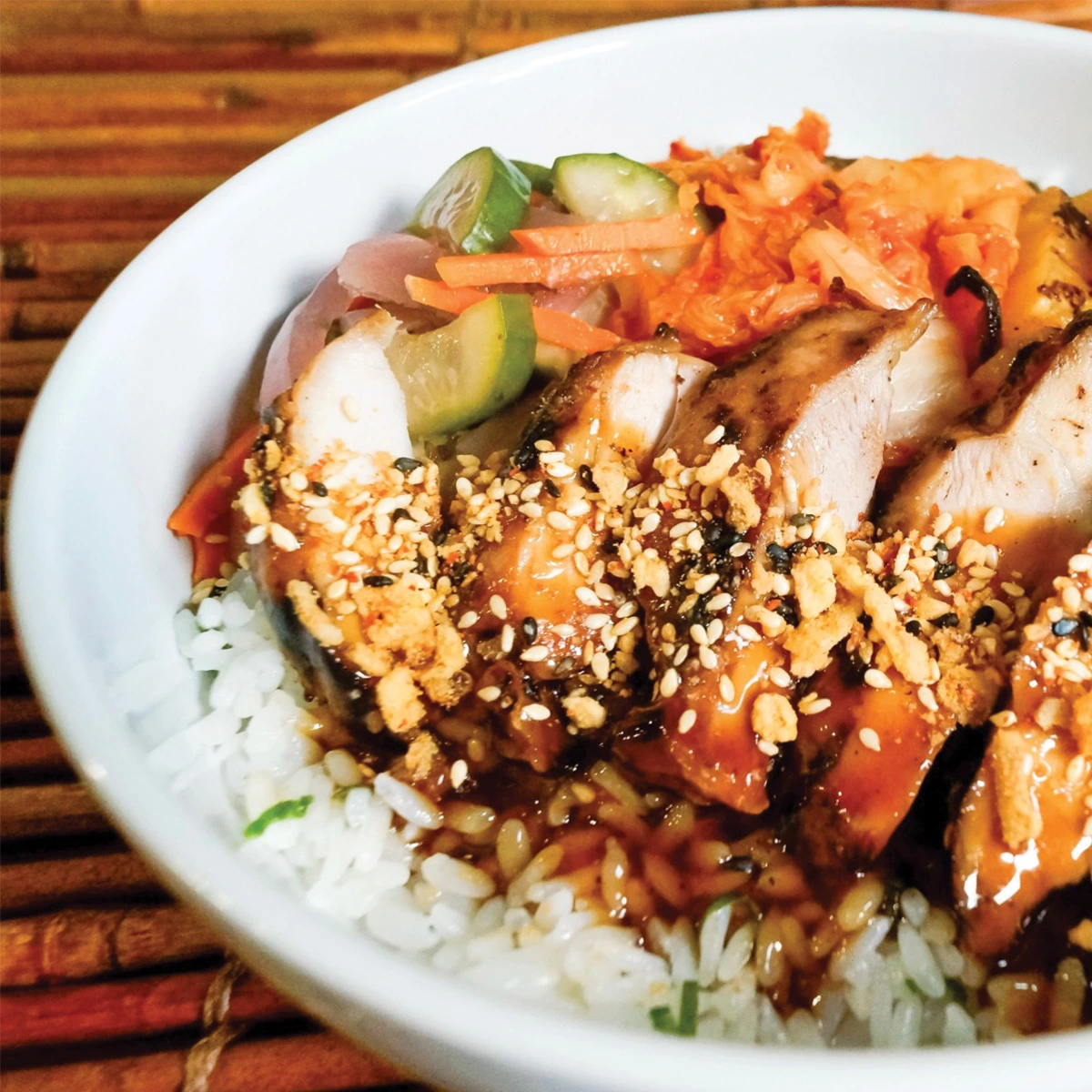 A bowl of rice topped with sliced glazed chicken, kimchi, pickled vegetables, and sprinkled with sesame seeds, on a wooden surface.