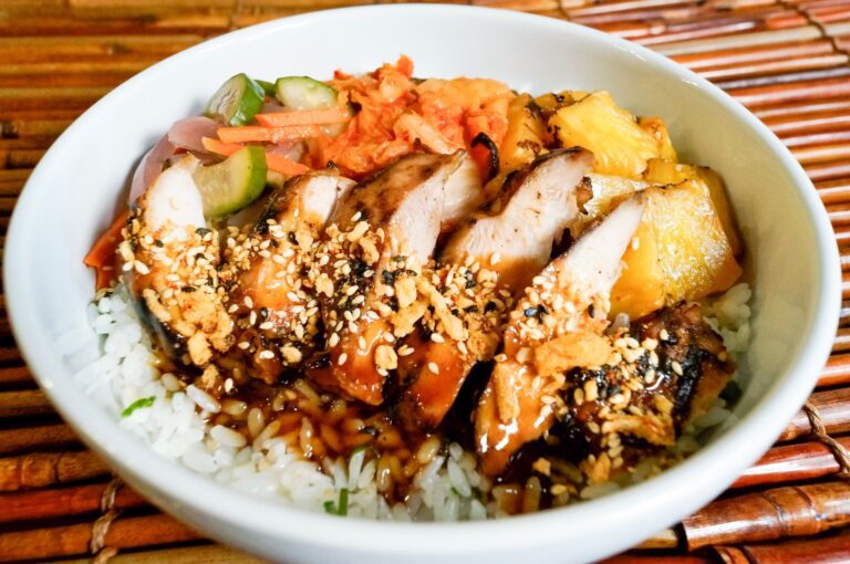 A white bowl of rice topped with sliced grilled chicken, sesame seeds, pickled vegetables, kimchi, and pineapple on a bamboo mat.