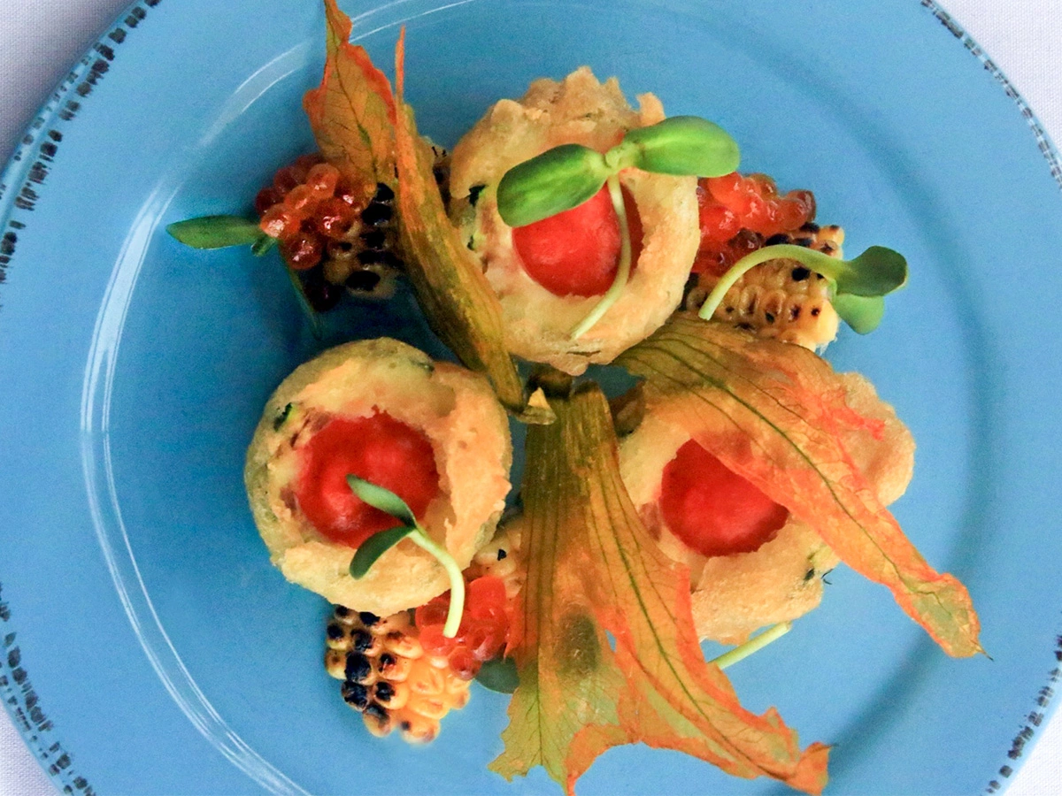 Three zucchini blossom fritters with red filling, grilled corn, salmon roe, microgreens, and crisp garnish on a blue plate.