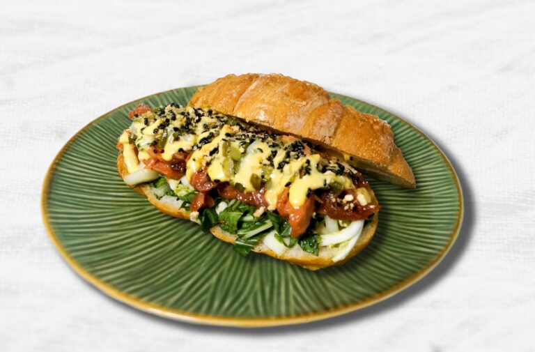 A croissant sandwich filled with greens, sliced egg, tomato, sauce, and a sprinkle of black sesame seeds, served on a green plate.