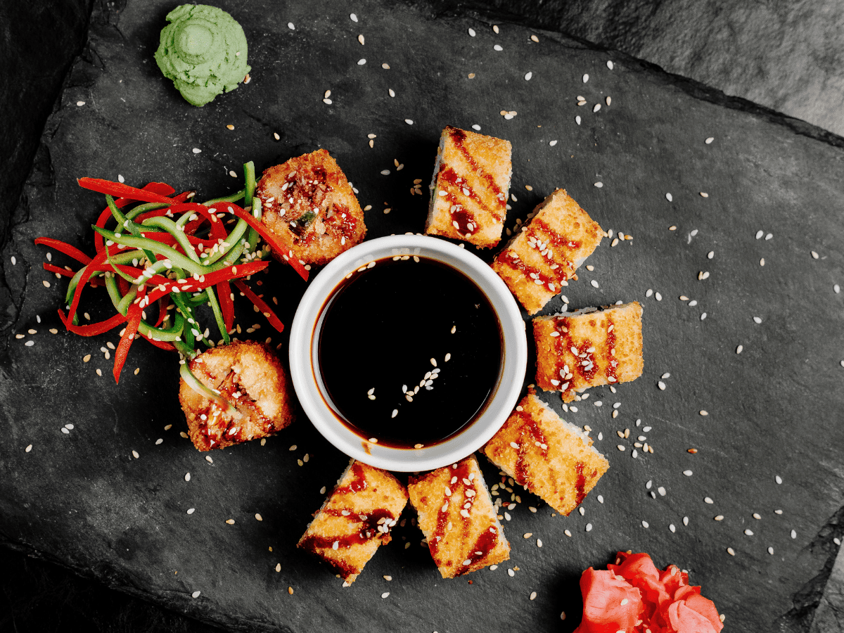 Eight pieces of sushi arranged around a bowl of soy sauce, garnished with sesame seeds, wasabi, red pickled ginger, and sliced vegetables on a black slate platter.