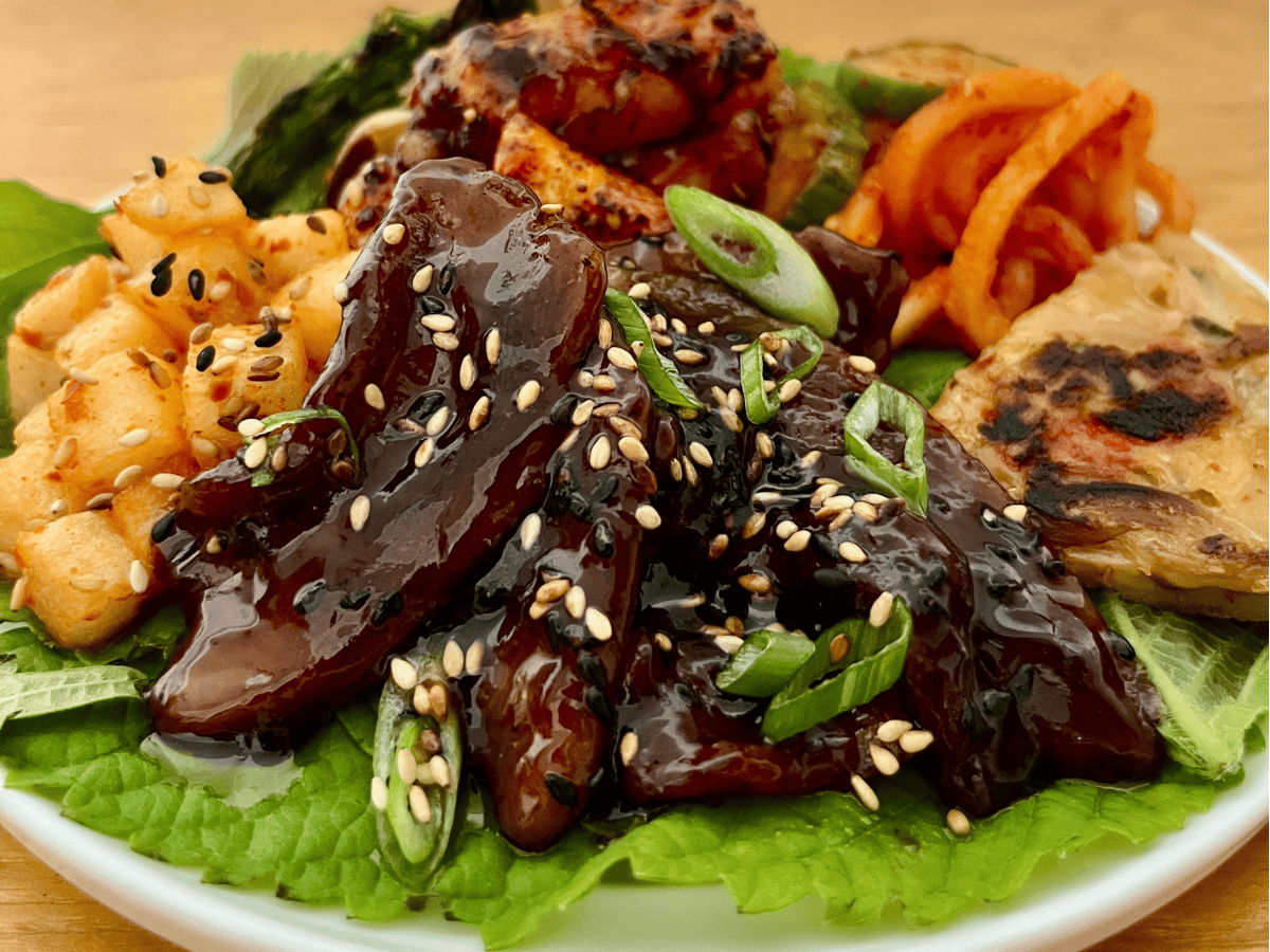 A plate with glazed beef, kimchi, pancakes, tofu, and vegetables, garnished with green onions and sesame seeds on a bed of lettuce.