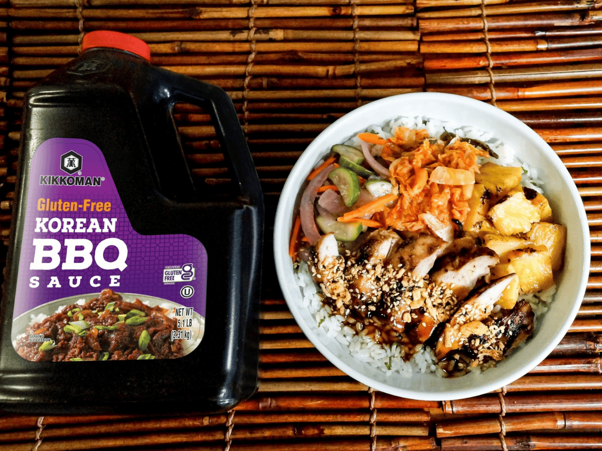 A large bottle of Kikkoman gluten-free Korean BBQ sauce next to a rice bowl with grilled chicken, pineapple, vegetables, kimchi, and sesame seeds, all on a bamboo surface.
