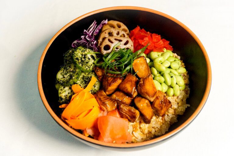 A bowl with rice, glazed tofu, edamame, cucumber, pickled ginger, shredded carrot, lotus root, red cabbage, and diced red pepper, garnished with sliced green onion.