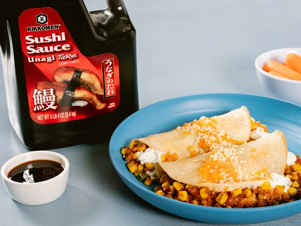 Plate of corn and cheese tacos with white sauce, next to a small dish of soy sauce, sushi sauce bottle, and a bowl of baby carrots on a light blue surface.