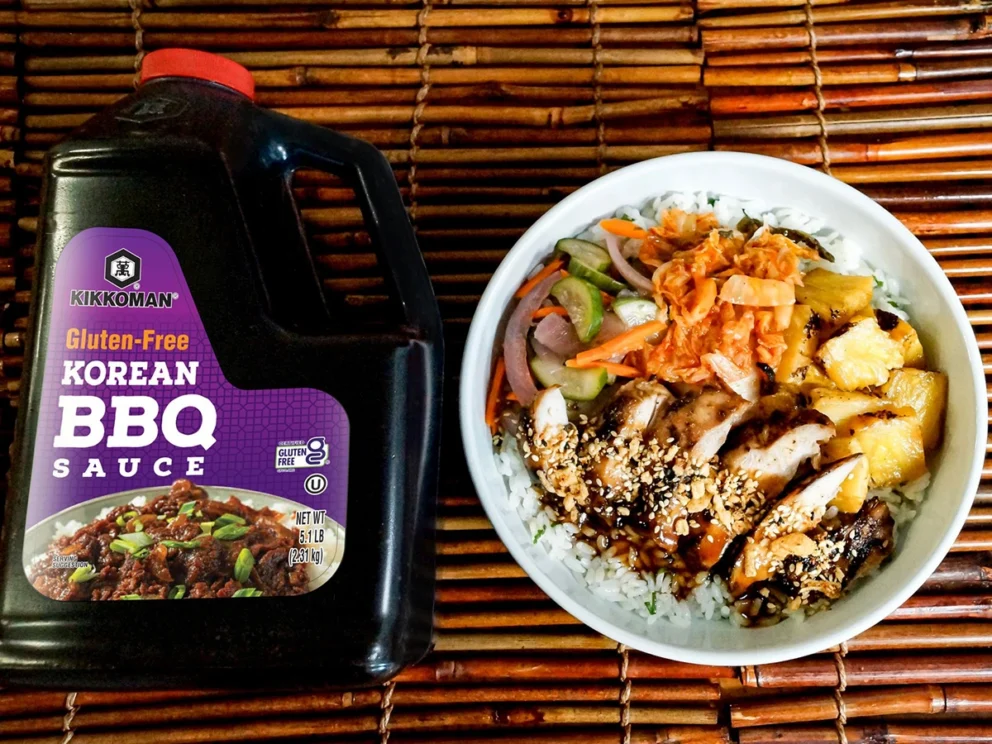 A bottle of Kikkoman gluten-free Korean BBQ sauce next to a rice bowl topped with grilled chicken, vegetables, kimchi, pineapple, and sesame seeds on a bamboo mat.