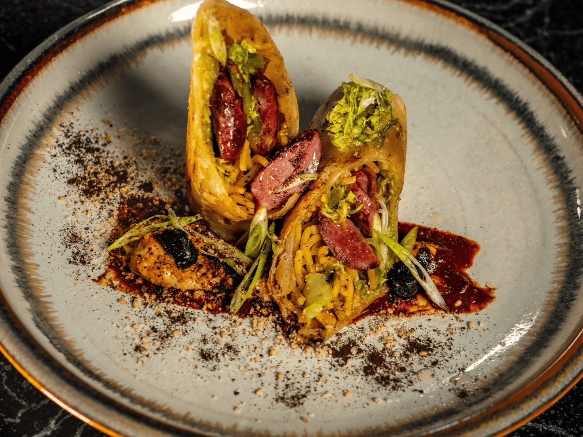 A plated dish featuring a sliced wrap filled with sausage, noodles, and greens, garnished with sauce, greens, and seasoning on a ceramic plate.